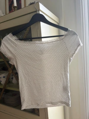 Vit prickig topp med spetsdetalj - Söt vit topp med små mörkblåa prickar och spetskant vid urringningen. Modellen är offshoulder med korta ärmar och har en feminin, figurnära passform. Perfekt för dig som gillar en enkel men ändå gullig stil. Har tyvärr lite missfärgningar under armarna från deodorant men det är inget som syns om man inte kollar noga.