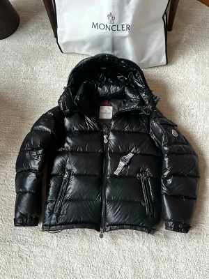 Svart dunjacka från Moncler - Snygg svart dunjacka från Moncler med glansig finish, avtagbar huva och två dragkedjefickor framtill. Jackan har Moncler-logga på ärmen och quiltad design som ger en puffig look. Perfekt nu inför kalla vinterdagar och riktigt trendig stil. Om det är något du undrar så ställ gärna frågor. Säljer den då den inte passar mig längre… Storleken är 1 vilket motsvarar ungefär S men passar också XS. 