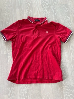 Röd Fred Perry pikétröja - Röd pikétröja från Fred Perry med klassisk krage och knappslå. Vita och marinblå ränder på krage och ärmslut samt broderad logga på bröstet. Tillverkad i bomull och har korta ärmar för en sportig look. Bara att höra av sig vid minsta lilla fundering!