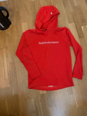hoodie från Peak Performance - Snygg orange hoodie från Peak Performance med vit logga tryckt på bröstet. Hoodien har långa ärmar, huva och är gjord i ett mjukt material som känns skönt mot huden. Perfekt för en avslappnad och sportig stil.