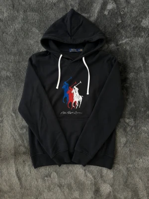 Svart hoodie Polo Ralph Lauren - Snygg svart hoodie från Polo Ralph Lauren med stor huva och vita dragsnören. På bröstet finns ett tryck med tre polospelare i blått, rött och vitt samt Polo Ralph Lauren-loggan under. Klassisk känguruficka framtill och ribbade muddar.
