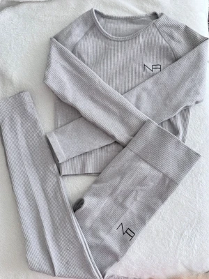 Grått ribbat loungewear-set - Mysigt tvådelat loungewear-set i ljusgrå ribbad trikå. Setet består av en långärmad tröja med rund hals och matchande leggings. Båda delarna har en diskret logga tryckt framtill. Perfekt för chill dagar hemma.