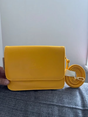 Gul axelväska från David Jones - Snygg gul axelväska från David Jones i slätt konstläder. Väskan har ett stilrent, rektangulärt format med lock och justerbar axelrem. Perfekt för dig som vill ha en färgklick till outfiten och gillar praktiska fack. Märkesdetalj på framsidan och sidan. Mått 20x16x6 cm