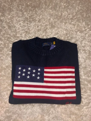 Mörkblå stickad tröja Polo Ralph Lauren - Snygg mörkblå stickad tröja från Polo Ralph Lauren med stor amerikansk flagga på bröstet. Tröjan har rund halsringning och ribbade muddar. Perfekt för dig som gillar klassisk preppy stil och vill sticka ut lite extra.