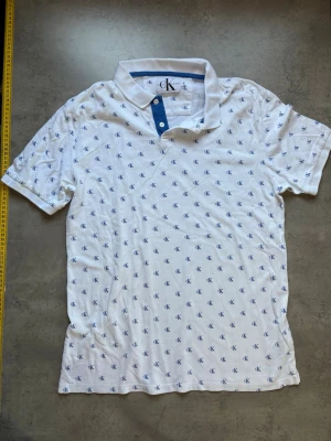 Vit piké från Calvin Klein Jeans - Vit pikétröja från Calvin Klein Jeans med blå cK-loggor över hela plagget. Tröjan har klassisk krage, två knappar framtill och korta ärmar. Perfekt för en clean och stilren look.