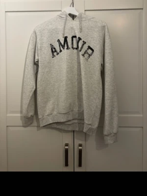 Ljusgrå hoodie med tryck - Säljer en ljusgrå hoodie med svart texten 'AMOUR' i ett coolt, lite slitet tryck över bröstet. Hoodien har huva med dragsko och ribbade muddar vid ärmar och nederkant. Perfekt för en avslappnad och trendig look.