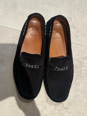 Svarta mockaloafers från HUGO BOSS - Snygga svarta loafers från BOSS i mjuk mocka med dekorativ kedjedetalj framtill. Skorna har klassisk form, bruna innersulor och räfflad gummisula för extra grepp. Tillverkade i Italien och perfekta för en stilren look.