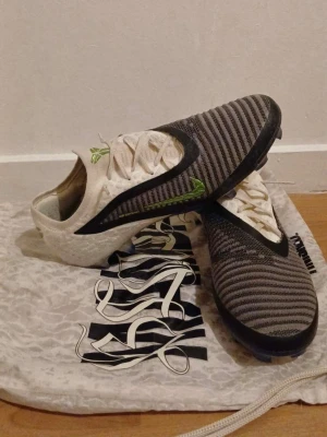 Nike Phontom fotbollsskor  - Säljer ett par fotbollsskor med svart och grått randigt mönster på ovandelen och vita partier med gröna detaljer. Skorna har snörning och låg profil, samt en sula anpassad för spel på konstgräs. Materialet ser ut att vara syntetiskt och designen är modern.