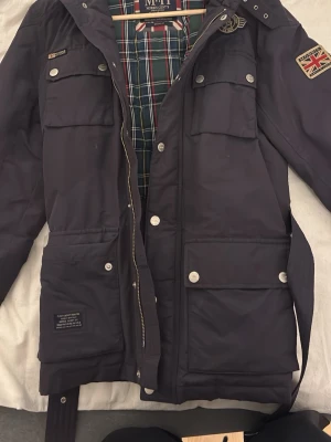 Mörkblå parkas från Morris Gents - Snygg mörkblå parkas från Morris Gents med rutigt innerfoder, fyra stora fickor framtill och dekorativa märken på bröst och ärm. Jackan dragkedja och knappar samt ett bälte i midjan för extra passform.