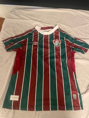 Randig Fluminense matchtröja från Umbro - Säljer en officiell Fluminense matchtröja från Umbro i storlek M. Tröjan har vertikala ränder i vinrött, grönt och vitt, med klubbmärke på bröstet och vit krage. Perfekt för fotbollsfans som vill sticka ut på planen eller läktaren.