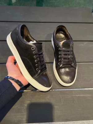Lanvin - Snygga svarta sneakers med lågt skaft i en mix av mocka och glansigt läder. Skorna har svarta snören, metallfärgade öljetter och en vit, tjock sula som ger en clean kontrast. Perfekta för dig som gillar stilrena och klassiska sneakers.