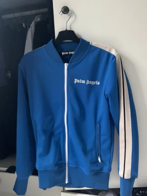 Blå ziptröja från Palm Angels - Snygg blå ziptröja från Palm Angels med vita detaljer och vita ränder längs ärmarna. Tröjan är i helt ok skick och är storlek XS 