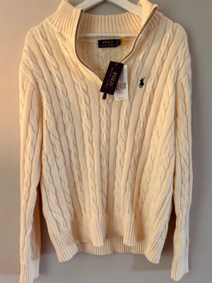 Halfzip Beige kabelstickad tröja från Polo Ralph Lauren - Kabelstickad tröja i Beige/Cream färg från Polo Ralph Lauren med klassisk marinblå logga på bröstet. Tröjan är i nyskick med tags på. Skick 10/10 utan defekter.