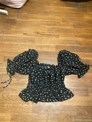 Svart blommig blus med puffärmar, strl S - Säljer en supersöt svart blus med små vita blommor och puffiga korta ärmar. Från H&M i storlek Small. Den har smockad midja och volang nedtill samt knytband vid ärmsluten. Säljes pga för liten🤗