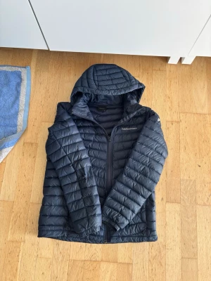 Svart dunjacka Peak Performance  - Snygg svart dunjacka från Peak Performance med huva och dragkedja framtill. Jackan har quiltad design och är perfekt för kalla vinterdagar. Lätt och smidig att bära, passar dig som vill ha både stil och funktion.