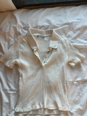 Ribbad vit polotopp från Zara - Säljer en ribbad vit polotopp från Zara med korta ärmar och klassisk krage. Toppen har knappar framtill. Storlek M från Zara. 