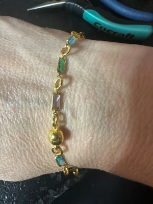 Guldigt armband med färgade pärlor - Snyggt armband i guldfärgad metall med rektangulära pärlor i olika färger som blått, grönt, lila och rött. Armbandet har små guldfärgade länkar mellan pärlorna och en rund guldfärgad detalj. Perfekt för dig som gillar färg och unika accessoarer.