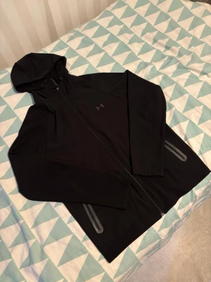 Svart träningsset från Under Armour - Snyggt svart träningsset från Under Armour med matchande hoodie och byxor. Hoodien har dragkedja, huva med dragsko och två fickor med dragkedja. Byxorna har resår i midjan och logga på benet. Perfekt för gymmet eller löprundan.