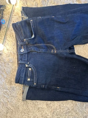Mörkblå raka jeans med rå kant - Säljer ett par mörkblå jeans med raka ben och rå kant nedtill. Jeansen har klassisk femficksdesign, silverfärgade nitar och kontrastsömmar. Materialet är denim i bomull och passformen är avslappnad. Perfekta för en cool och enkel stil.