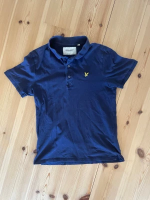 Marinblå piké från Lyle & Scott - Snygg marinblå pikétröja från Lyle & Scott i ekologisk bomull. Klassisk krage med knappar och den ikoniska gula loggan broderad på bröstet. Perfekt till jeans eller shorts för en clean och stilren look.