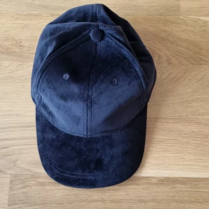 Svart keps från Gina Tricot - En svart sammets keps från Gina Tricot tillverkad av 100% polyester. Kepsen har en justerbar rem på baksidan för att passa olika huvudstorlekar.