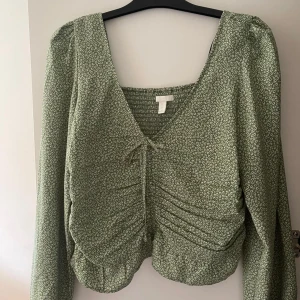 Grön blommig blus - Säljer denna grön blommiga blus från H&M med långa ärmar och en v-ringad hals. Blusen har en knytning framtill och rynkade detaljer på framsidan. Endast använd en gång och är i storlek L. ✨🌸