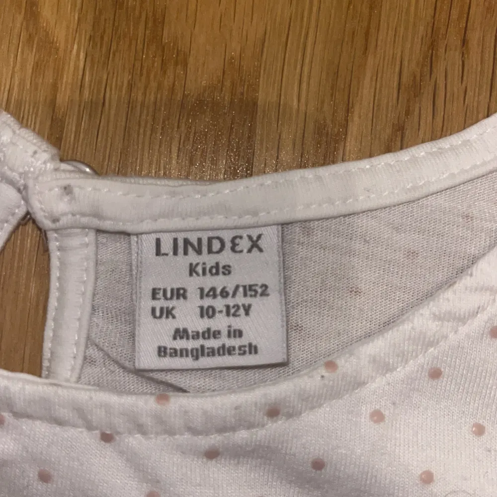En vit topp från Lindex med små bega prickar. Toppen har korta ärmar med volangdetaljer och en knappöppning i nacken.. Yläosat.