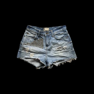 Blå jeansshorts med nitar - Ett par blå jeansshorts med slitna detaljer och nitar på framsidan. Shortsen har en rå kant och är lågmidjade. De har fem fickor och en knappgylf.