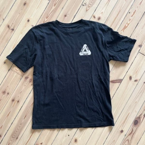 Svart t-shirt från Palace - En svart t-shirt från Palace med ett tryckt triangulärt mönster på bröstet och ett större liknande mönster på ryggen i färgerna röd, blå och vit. T-shirten har korta ärmar och en normal passform.