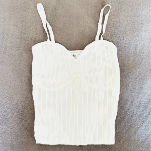 Vit topp Bershka XS - Färg: vit.  Strl: XS.  Snyggt linne från Bershka. Ribbat tyg, snygga dekaner av framsydda kupor framtill. Reglerbara band. Skönt stretchmaterial! OBS - Helt ny & oanvänd!