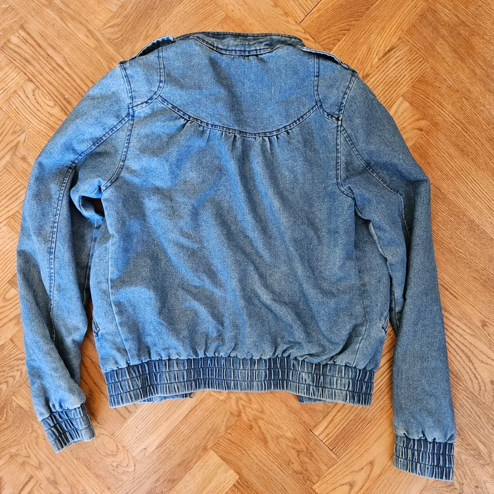 En jeansblå jacka med långa ärmar och elastiska muddar vid ärmslut och nederkant. Jackan har en ståkrage med knapp och två framfickor. Materialet är jeans och insidan är fodrad med ett mörkt tyg.. Jackor.