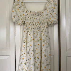 Blommig klänning med puffärmar - En vacker klänning med blommönster i gult och grönt på en vit bakgrund. Klänningen har puffärmar och en smockad överdel som ger en bekväm passform. Den är tillverkad i ett lätt och luftigt material, perfekt för varma dagar. Viskos och bomull. 