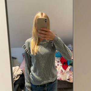 Grå stickad tröja med volangdetaljer - Säljer en superfin grå tröja med volangdetaljer på axlarna. Den är perfekt för höst och vinter, och är både varm och stilren. Tröjan har långa ärmar och en normal passform.  💖