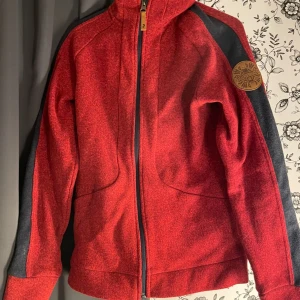 Röd fleecejacka med svarta detaljer - Säljer en snygg och bekväm röd fleecejacka med svarta detaljer på ärmarna. Jackan har en dragkedja framtill och en hög krage för extra värme. Perfekt för kyliga dagar!