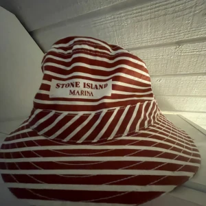 Röd- och vitrandig hatt från Stone Island - Säljer en snygg röd- och vitrandig hatt från Stone Island. Perfekt för sommaren med sitt marina tema. Hatten är i bomull och har en avslappnad passform. Märket 'Stone Island Marina' är broderat på framsidan. Passar både för en dag på stranden eller en sommarfest.