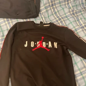 Svart Jordan tröja med röd och vit text - Säljer en snygg svart Jordan tröja i storlek L. Tröjan har långa ärmar och texten 'Jordan' i rött och vitt på bröstet samt längs ärmarna. Den är gjord av Dri-Fit material vilket gör den perfekt för träning eller vardagsbruk. Tröjan är i bra skick och redo för en ny ägare!