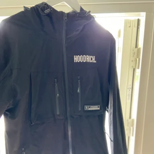 Svart vindjacka från Hoodrich - Säljer en svart vindjacka från Hoodrich i storlek L. Jackan har en dragkedja framtill, två bröstfickor med dragkedja och en liten logga på vänster sida. Den är perfekt för vår och höst, och har en normal passform. Materialet känns slitstarkt och den har en huva för extra skydd mot väder och vind.