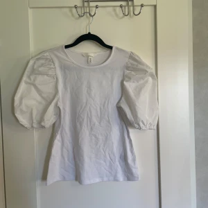 Vit topp med puffärmar - Säljer en superfin vit topp med puffärmar. Den är i storlek S och har en normal passform. Perfekt för både vardag och fest! Toppen är i nyskick och gjord av bomull, vilket gör den både bekväm och stilren. Passar bra till både jeans och kjol.