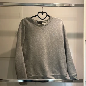 Grå sweatshirt från Polo Ralph Lauren - Säljer en snygg och bekväm grå sweatshirt från Polo Ralph Lauren. Tröjan har en enkel design med en liten broderad logga på bröstet. Perfekt för en avslappnad och stilren look. Passar bra till både vardag och träning.