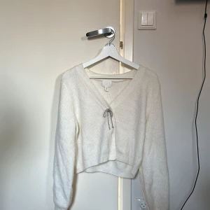 Stickad kofta med glitter detalj - Vit kofta med en rosett av glitter. Kan knäppas upp, men knapparna är svåra att få upp. Rekommenderar att inte knäppa upp då trådarna inte sitter så bra