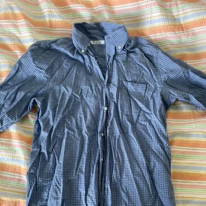 Blå rutig skjorta från Loro Piana - En blå rutig skjorta från Loro Piana, tillverkad i Italien. Skjortan har långa ärmar och knappar framtill samt en bröstficka.