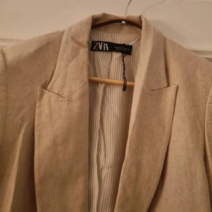 Beige kavaj från Zara - En stilren beige kavaj från Zara. Kavajen har en klassisk design med två fickor framtill och en enkel knäppning. Den är tillverkad i linne material som gör den perfekt för både vår och höst.