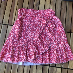 Röd blommig kjol från Shein - En rödblommig kjol från Shein med volangdetaljer och knytband. Kjolen är kort och har ett elastiskt midjeband för en bekväm passform. Tillverkad av 65% bomull och 35% polyester.