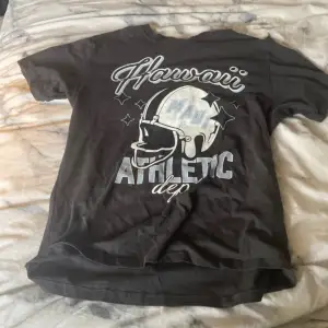 En grå t-shirt med korta ärmar och ett tryck på framsidan som visar en hjälm och texten 'Hawaii Athletic dep'. T-shirten är i storlek dam xxs, texten har tappat lite men det ser man på tredje bilden