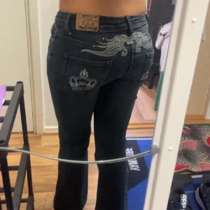Svarta jeans med broderi - Ett par svarta jeans med broderade detaljer på bakfickorna och ovanför. Jeansen har en bootcut-stil och är lågmidjade. Dom är lite sönder vid där man sätter runt bältet 