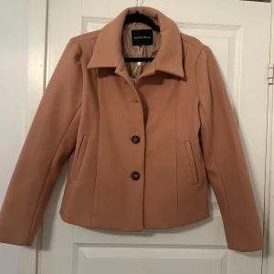 Beige kappa från Rut&Circle - En elegant beige kappa från Rut&Circle. Jackan har långa ärmar, två knappar framtill och två fickor. Den är fodrad och har en klassisk krage.