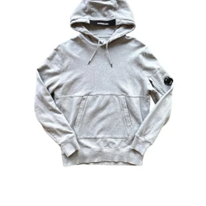CP COMPANY - En bekväm grå hoodie med en ficka framtill och en detalj på vänster ärm. Hoodien har en justerbar huva med dragsko och ribbstickade muddar vid ärmslut och nederkant. CP Company. 