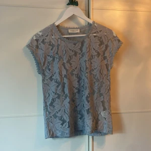 Grå spetsblus från Rosemunde - En elegant blågrå blus från Rosemunde i genombruten spets. Blusen har korta ärmar och en rund halsringning med spetsdetaljer.