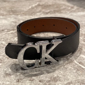 Svart skärp med CK-spänne - Ett elegant svart skärp med ett stort metallspänne i form av bokstäverna CK. Skärpet är tillverkat av skinn och har en slät yta.
