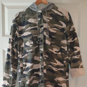 Armejacka stl. 34 - Tuff huvjacka av märket Divided. Gråmelerad trikå luva med snören. Två bröstfickor och två sidfickor. Knäpoes med knappar. 100% bomull.Tvättas i 40 grader. Fint skick. Priset är inklusive frakt inom Sverige 